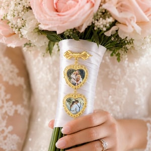 Pu&ograve; includere: Un ciondolo in metallo dorato per bouquet da sposa con due cornici a forma di cuore contenenti fotografie, sospeso a una spilla decorativa a forma di fiocco. Il ciondolo &egrave; fissato a un manico di bouquet avvolto in raso bianco con rose rosa.