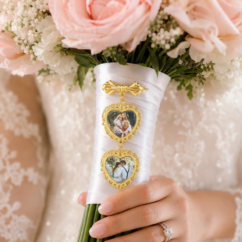 Pu&ograve; includere: Un ciondolo in metallo dorato per bouquet da sposa con due cornici a forma di cuore contenenti fotografie, sospeso a una spilla decorativa a forma di fiocco. Il ciondolo &egrave; fissato a un manico di bouquet avvolto in raso bianco con rose rosa.