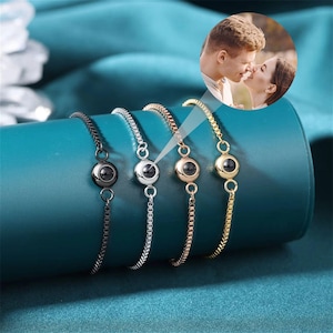 以下が含まれることがあります： 小さな黒い丸いチャームが付いた銀色のブレスレットが4つ。各チャームには、特定の角度から見ると隠された画像が明らかになる、小さな透明な丸い窓があります。