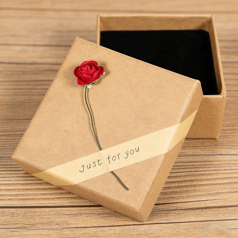 May include: A square brown cardboard gift box with a black velvet interior lining. The lid features a diagonal cream-colored ribbon printed with the text Just for you and a small artificial red rose attached to the stem of the ribbon.
