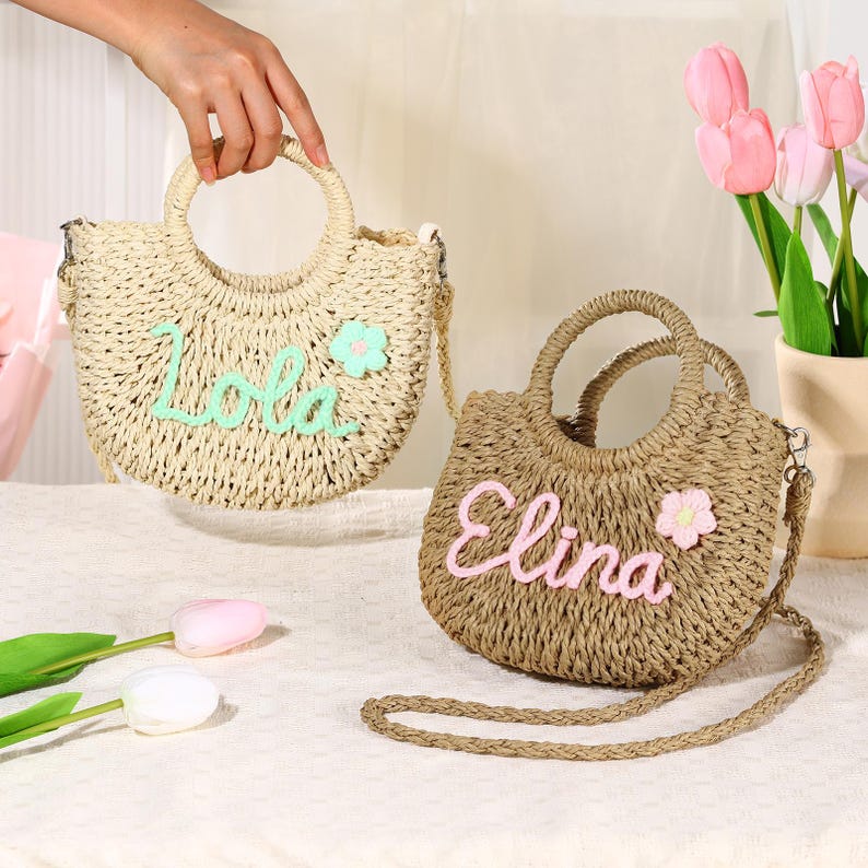 May include: Two straw tote bags with personalized names. The bag on the left is light brown with a green flower and the name "Lola" embroidered on it. The bag on the right is a darker brown with a pink flower and the name "Elina" embroidered on it.