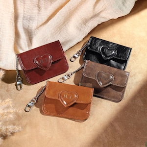 May include: Four small leather wallets in shades of brown and black, each with a heart-shaped accent and a chain. The wallets have a flap closure and the initials "H.J." are visible on the heart. The wallets are arranged on a neutral surface.