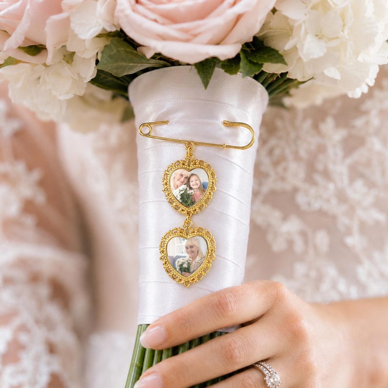 Pu&ograve; includere: Una spilla da balia color oro con due cornici a forma di cuore dai bordi smerlati. Ogni cornice contiene una piccola fotografia di persone. Il ciondolo &egrave; fissato al manico di un bouquet avvolto in raso bianco, sotto delle rose rosa pallido.