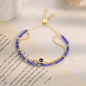 May include: A gold adjustable bracelet with a blue crystal bead chain and a small gold charm with a black eye design. The bracelet has a sliding clasp.