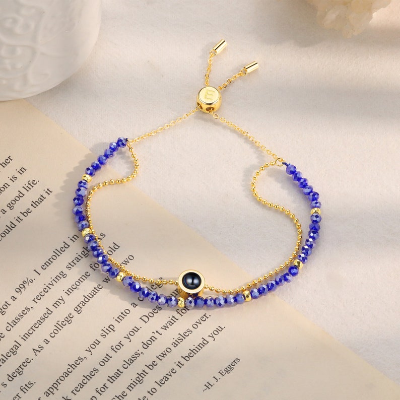 May include: A gold adjustable bracelet with a blue crystal bead chain and a small gold charm with a black eye design. The bracelet has a sliding clasp.