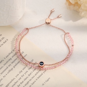 May include: A rose gold bracelet with a pink stone charm and a small metal charm with the letter "N" engraved on it. The bracelet is adjustable and has a sliding clasp.