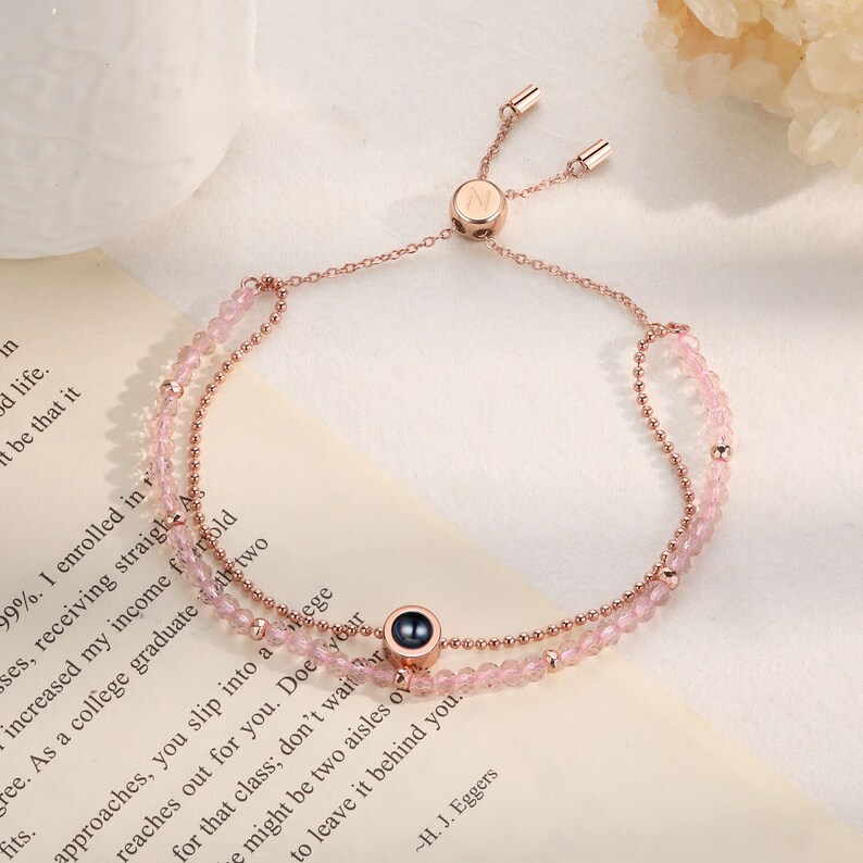 May include: A rose gold bracelet with a pink stone charm and a small metal charm with the letter "N" engraved on it. The bracelet is adjustable and has a sliding clasp.