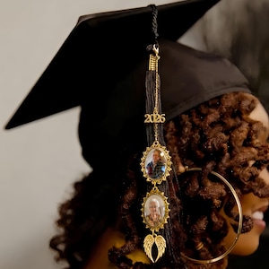 May include: A black graduation cap featuring a black tassel adorned with a gold-toned charm displaying the year 2026, two oval-shaped gold-toned photo frames containing portraits, and a pair of small gold-toned angel wings at the bottom.