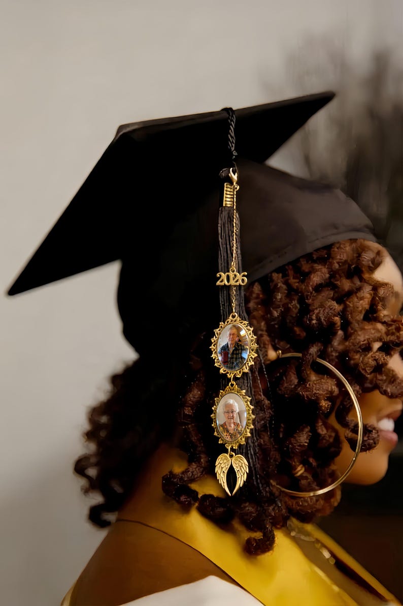 May include: A black graduation cap featuring a black tassel adorned with a gold-toned charm displaying the year 2026, two oval-shaped gold-toned photo frames containing portraits, and a pair of small gold-toned angel wings at the bottom.