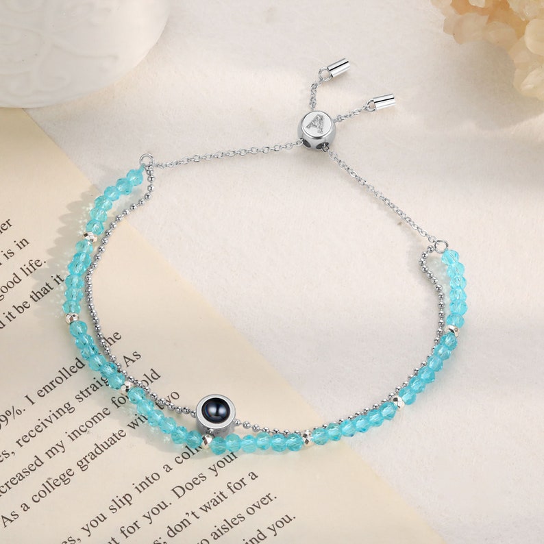 May include: A silver chain bracelet with a sliding clasp and two rows of light blue beads. The bracelet has a small black and silver charm with a round black stone in the center.