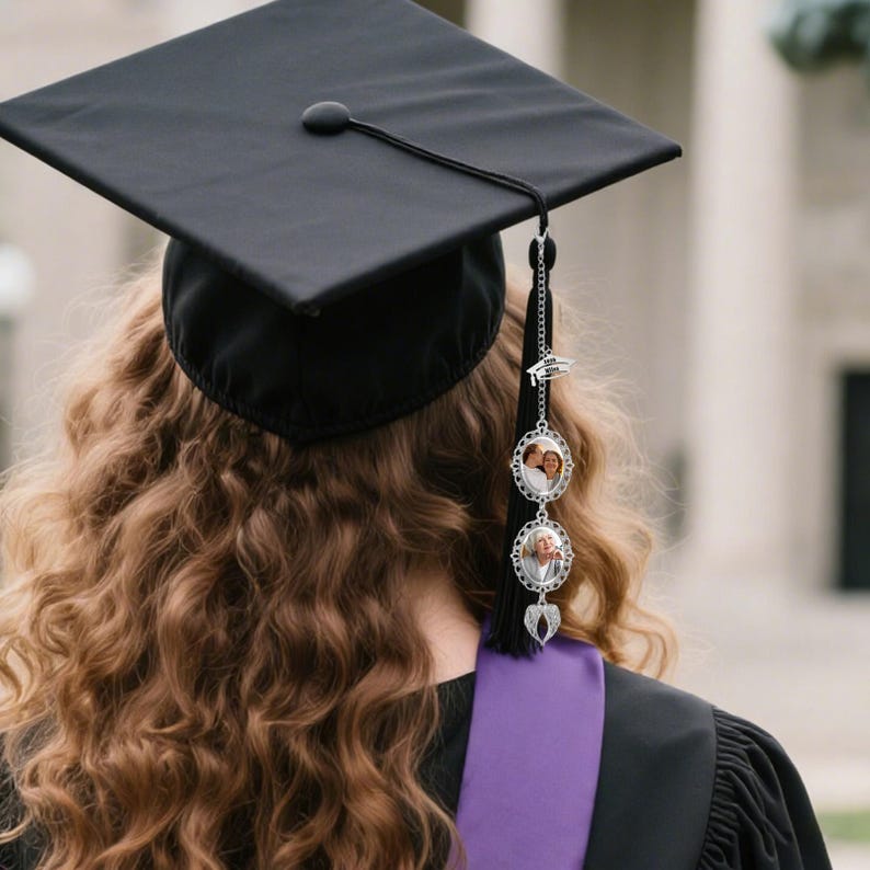 May include: A black graduation cap with a black tassel and a silver-toned metal charm attachment. The charm features a small graduation cap icon, two oval frames containing photographs, and a pair of silver angel wings at the bottom of the chain.