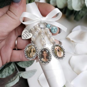 May include: A white ribbon-wrapped bouquet charm featuring three oval silver-toned photo frames, a heart-shaped pendant engraved with the text you are always in my heart, white faux pearl beads, and a single light blue bead tied with a white bow.