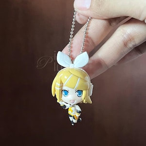 Może przedstawiać: Mała figurka w stylu anime z żółtymi włosami i niebieskimi oczami, przymocowana do srebrnego łańcuszka. Figurka ma na sobie biało-żółty strój. Tło to rozmyta brązowa powierzchnia. Tekst na dole brzmi: (only on etsy https://www.etsy.com/shop/pinkdayplush)