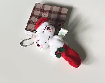 May include: A white plush Christmas ornament with a Santa hat and a red stocking. The ornament has a silver chain and is next to a small notebook with a plaid pattern.