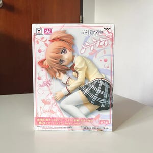 Pode incluir: Uma figura de anime em caixa de um personagem com cabelo cor de pêssego, vestindo um top amarelo, saia xadrez e meias altas brancas. A caixa apresenta texto japonês e um design floral. A figura é da série "Madoka Magica".