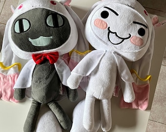 May include: Two plush dolls with white hooded outfits and long ears. One doll has a dark grey face with black oval eyes and a red bow tie. The other doll has a white face with pink cheeks and a smiling expression. Both dolls have white capes and pink accents.