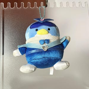 Puede incluir: Un peluche de pingüino en tonos azules, blancos y amarillos. El pingüino tiene un sombrero azul, una pajarita y una banda de cinta. El juguete está colgado de una pantalla de plástico transparente.