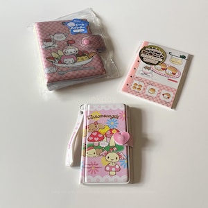 Puede incluir: Carpetas rosas y blancas estampadas y un pequeño cuaderno. Las carpetas presentan ilustraciones de dibujos animados de personajes y comida. Una carpeta tiene un cierre de botón y la otra un cierre en forma de corazón. El cuaderno tiene un borde rosa.