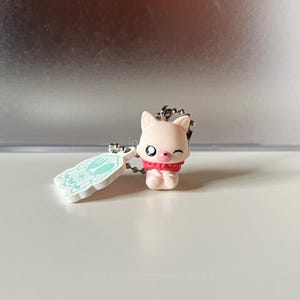 Könnte beinhalten: Ein hellrosa Tierfiguren-Schlüsselanhänger mit zwinkerndem Gesichtsausdruck und rotem Halsband. An der Metallkugelkette befindet sich ein weißer Acryl-Anhänger mit einem hellblauen Grafikdruck von Gabel, Löffel und Teller in einem gewellten Rahmen.
