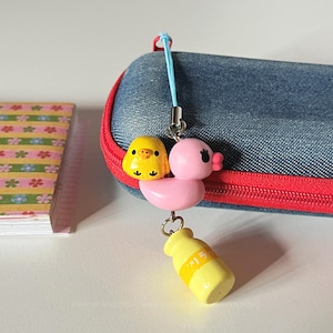 Pode incluir: Um charme caprichoso com um pintinho amarelo e um pato rosa, com um charme de garrafa amarela. O charme é preso a um estojo de lápis jeans azul com zíper vermelho. Um pequeno caderno floral é visível à esquerda.