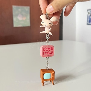Puede incluir: Un llavero con un conejito rosa claro, un bloque rosa con texto japonés y un pequeño televisor marrón. El conejito está en una pose dinámica. El televisor tiene una pantalla azul claro y cuatro patitas.