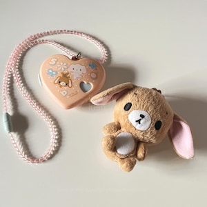 Puede incluir: Un colgante en forma de corazón melocotón con un collar de cuentas rosa, con personajes de dibujos animados y diseños florales. También hay un conejito de peluche marrón con orejas internas rosas y vientre blanco. Los artículos están sobre una superficie blanca.