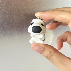 May include: A white plastic dog figurine keychain featuring a large black nose and black eyes. The small, cartoon-style puppy character has a smooth matte finish and is attached to a silver-toned metal ball chain for hanging as a decorative accessory.