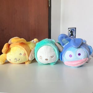 May include: Three plush toys in yellow, teal, and blue. Each plush has a round body and is wearing a hooded outfit. The yellow plush has a smiling face and pink cheeks. The teal plush has a white face and closed eyes. The blue plush has large eyes and a pink mouth.