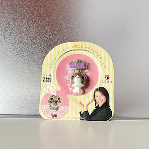 May include: A small brown and white Scottish Fold cat figurine keychain attached to a yellow and pink cardboard backing. The packaging features Japanese text, a photo of a real cat, and a graphic of a person with hands raised. The logo reads THE CAT.
