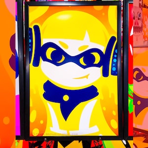 May include: A framed art print featuring a stylized yellow character with a blue mask, blue headphones, and a blue scarf against a bright orange and yellow background. The text SHOP.MELADRENALINE.COM is visible on the left side of the frame.
