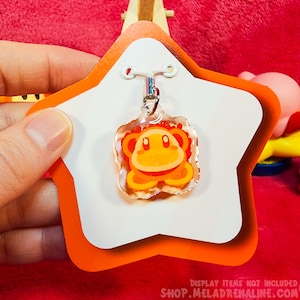 May include: A clear orange and yellow star-shaped keychain charm with a cartoon character inside. The charm is hanging from a silver chain and is displayed on a white star-shaped card with an orange border. The text "SHOP.MELADRENALINE.COM" is printed on the card.