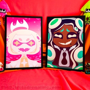 May include: Two framed prints of cartoon characters. The print on the left features a pink and white character with a crown and large eyes. The print on the right features a green and black character with large eyes and a smile.