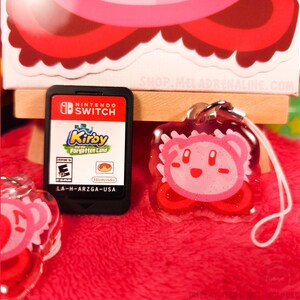 以下が含まれることがあります： 白い紐につけられた、ピンクと白のキラキラしたカービィのキャラクターチャーム。チャームはキーホルダーまたは携帯電話のチャームです。画像には、カービィと忘れられた星のNintendo Switchゲームカートリッジも表示されています。