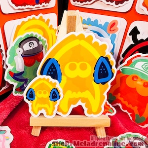 Può includere: Un set di adesivi di personaggi dei cartoni animati colorati che presentano una creatura gialla, arancione e verde con un cappello verde, una creatura gialla con gli occhi blu e una creatura gialla con una benda sull'occhio nera. Gli adesivi sono esposti su un cavalletto di legno.