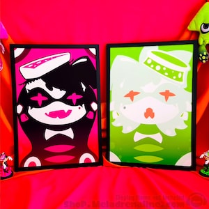 以下が含まれることがあります： 2つの額縁に入った漫画のキャラクターのプリント。左側のキャラクターはピンクと黒で、白い顔と黒い帽子をかぶっています。右側のキャラクターは緑と白で、白い顔と緑の帽子をかぶっています。