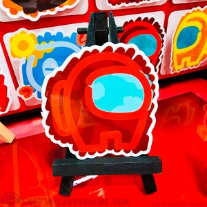 May include: Red Among Us character sticker with a white outline and a blue visor. The sticker is on a small black easel.