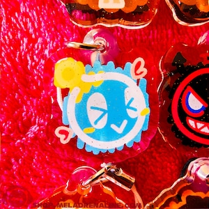 以下が含まれることがあります： 透明なプラスチック製のキーホルダーで、青と白の漫画のキャラクター、黄色の太陽、ピンクのハートが付いています。キャラクターは青い顔に白い目と頬に白いチェックマークが付いています。キーホルダーは透明なプラスチック製で、金色の金属製のリングが付いています。