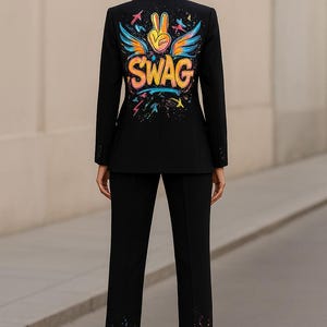 May include: A black blazer and matching trousers set. The back of the blazer features a colorful graphic print with the word SWAG, a peace sign, and wings. The bottom of the trousers features a colorful paint splatter design in blue, yellow, and red.