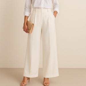 May include: A woman wearing a white button-down shirt with rolled-up sleeves and cream-colored wide-leg pants. She is holding a tan clutch and wearing beige heels. The outfit is a classic, elegant style.