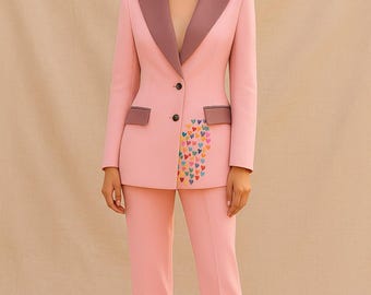 May include: A light pink suit with a blazer and matching trousers. The blazer has a contrasting mauve lapel and pocket trim, with a colorful heart design on the front. The suit is tailored and worn with nude heels.