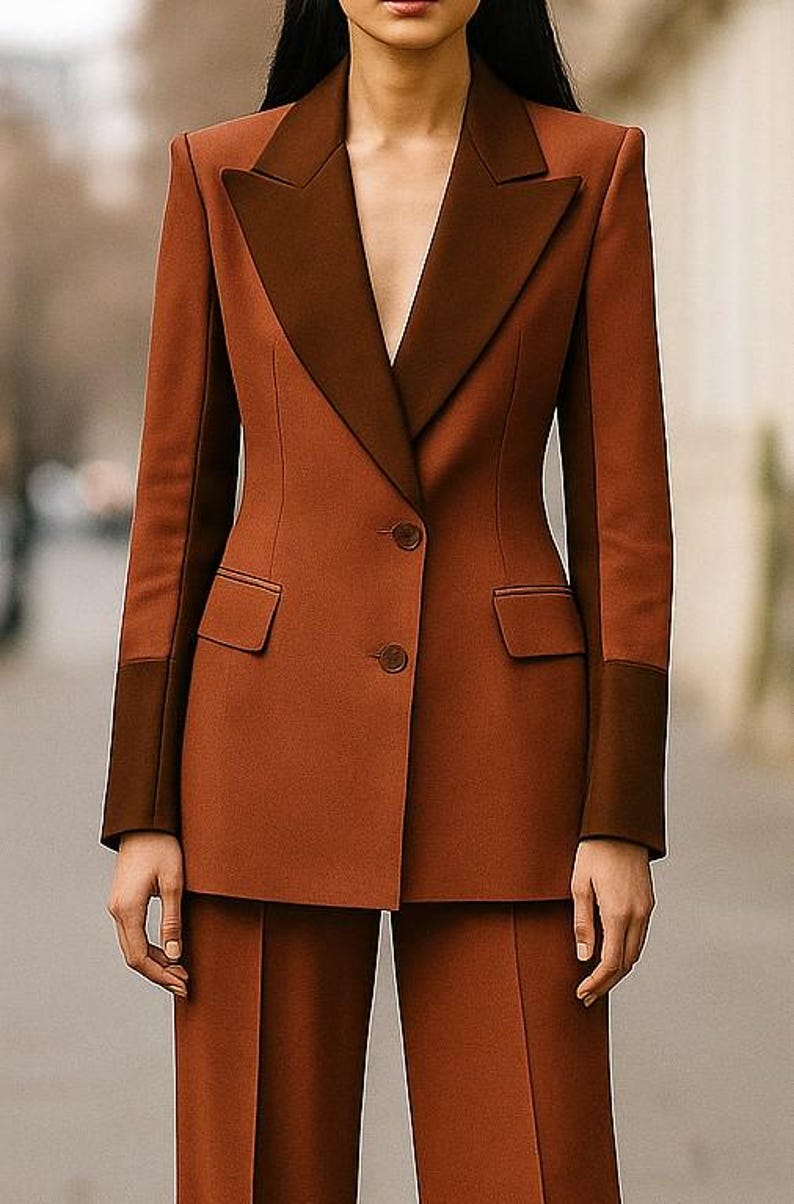 May include: A two-tone rust-colored suit featuring a tailored blazer and matching trousers. The blazer has a contrasting dark brown lapel and sleeve cuffs, with two front pockets and two buttons. The suit is a classic, elegant style.