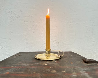May include: A lit candle in a brass candlestick on a dark wooden surface. The candle is yellow and the candlestick has a ring handle.