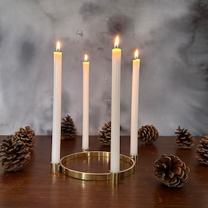 Könnte beinhalten: Ein Kerzenhalter aus Messing mit vier brennenden weißen Kerzen. Der Halter ist rund und hat ein schlichtes Design. Die Kerzen sind in einem Kreis um den Halter angeordnet.