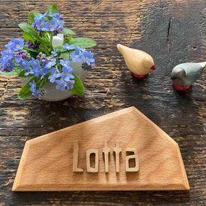 Może przedstawiać: Drewniany znak z wygrawerowanym imieniem "Lotta". Znak leży na drewnianym stole z małym bukietem niebieskich niezapominajek i dwiema drewnianymi figurkami ptaków.