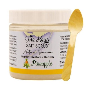 Puede incluir: Un tarro de "The Keys Salt Scrub" con aroma a pi&ntilde;a, con tapa blanca y etiqueta amarilla. Una cuchara de madera descansa junto al tarro. El producto es para el cuidado natural de la piel.