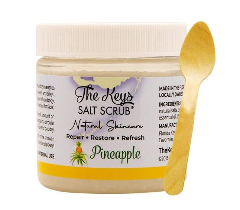 Puede incluir: Un tarro de "The Keys Salt Scrub" con aroma a pi&ntilde;a, con tapa blanca y etiqueta amarilla. Una cuchara de madera descansa junto al tarro. El producto es para el cuidado natural de la piel.