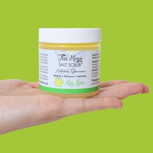 May include: A jar of The Keys Salt Scrub in Key Lime. The jar has a white lid and a label with text and a lime illustration. The product is held in a hand against a green background. The text on the label says "Natural Skincare".