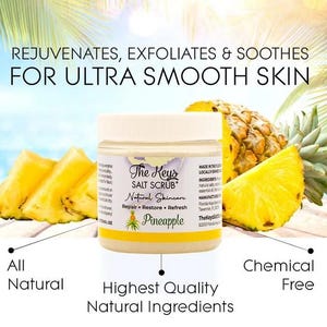 Puede incluir: Un tarro de exfoliante de sal The Keys con etiqueta "Pi&ntilde;a", rodeado de rodajas de pi&ntilde;a fresca. El texto dice "Rejuvenates, Exfoliates & Soothes For Ultra Smooth Skin." El texto adicional incluye "All Natural" y "Chemical Free."