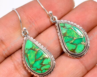 May include: A pair of silver dangle earrings featuring green turquoise stones. The stones have a mottled green colour with gold flecks. The earrings are teardrop shaped and set in a silver bezel.