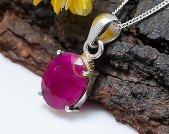May include: A silver pendant necklace with a large, oval-cut ruby gemstone. The ruby is a deep magenta colour, set in a silver bezel with four prongs. The chain is a delicate silver link. A yellow flower is in the background.